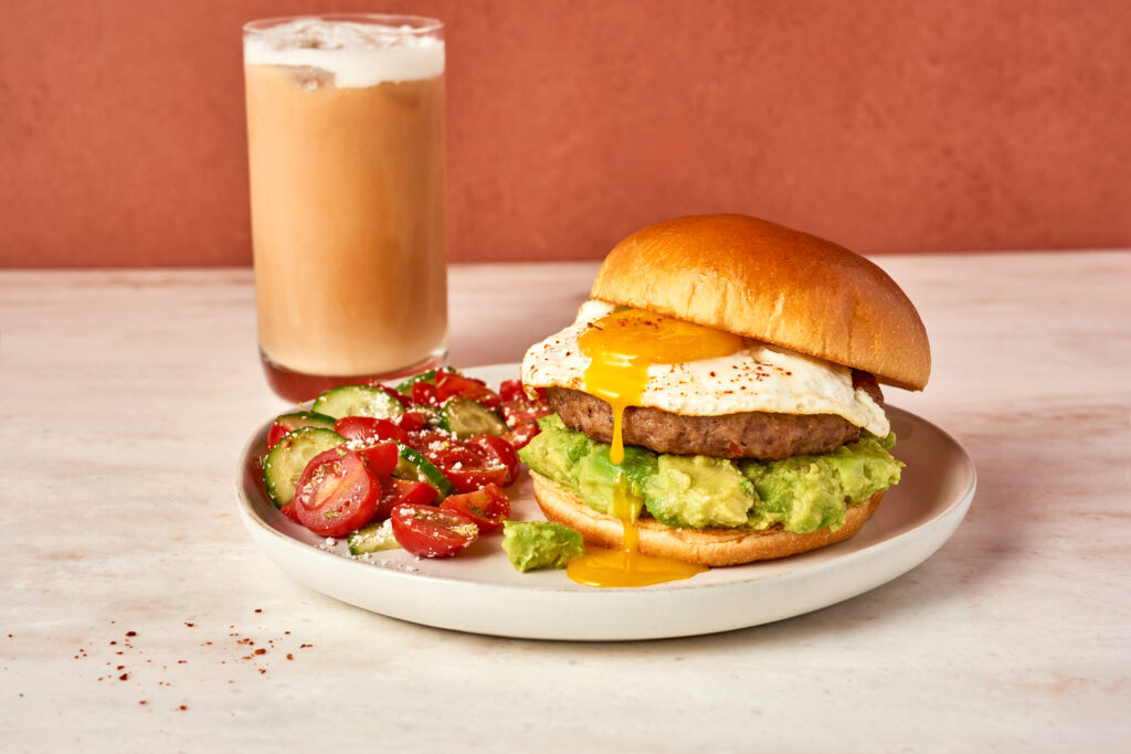 A breakfast sandwich on a plate with egg, sausage and avocado.