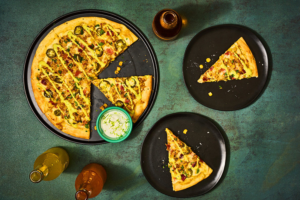 Overhead shot of pizza topped with street corn, accompanied by a few beverages
